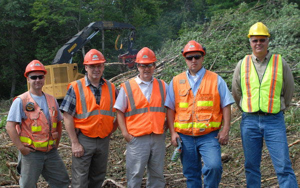 3 men in safety gear stand together on a job site in front of a Tigercat processor.