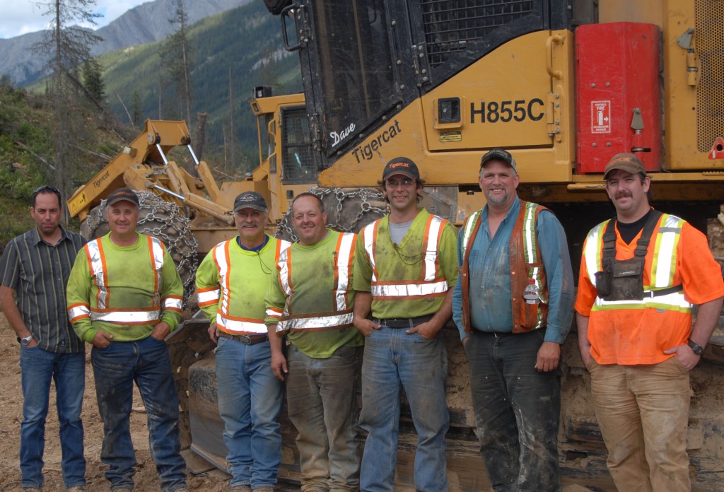 Dale Felhauer (tout à gauche), directeur de la succursale de Parker-Pacific à Cranbrook, avec les membres de l'équipe de Carl Larson’s Enterprise Ltd. (de gauche à droite) : Lance Larson, Ivar Larson, David Deveau, Jory Langridge, Bob Findlay et George Barbour. Le groupe pose devant une abatteuse-façonneuse et un débardeur Tigercat, sur un fond de paysage montagneux.