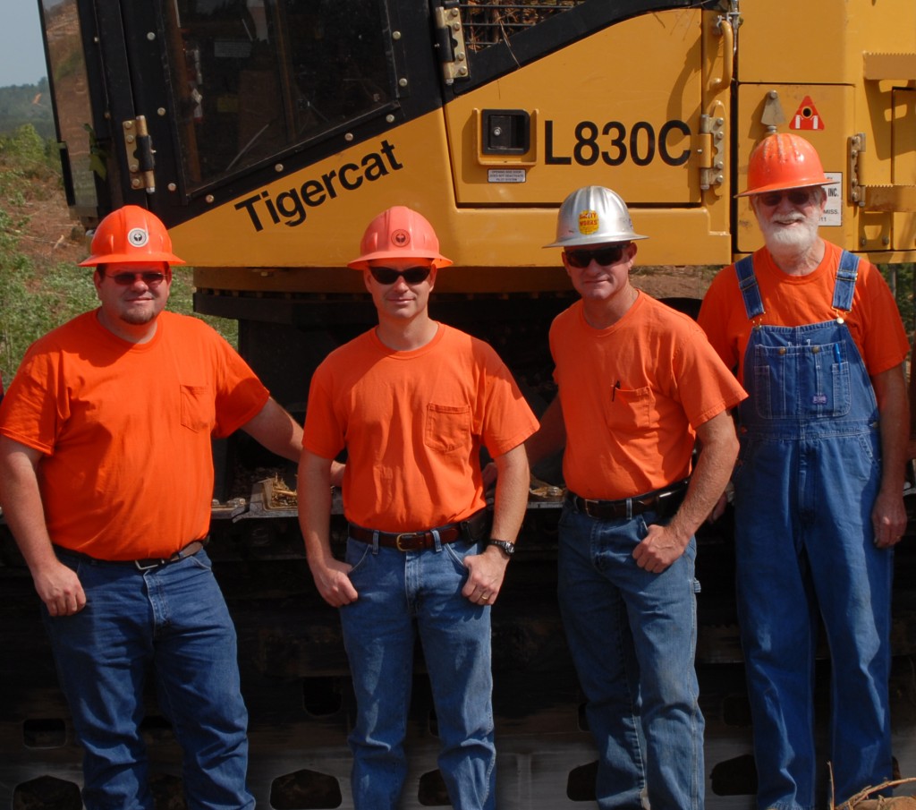 Derrick, Kevin, Tim and Sam O’Bryant stand together in matching orange t-shirts and blue jeans in front of their L830C track machine.