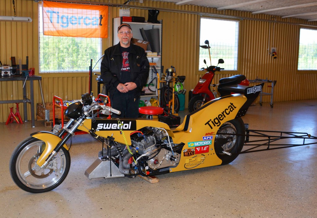 Mats standing with his drag bike inside a workshop, a Tigercat flag hangs in the window behind him.