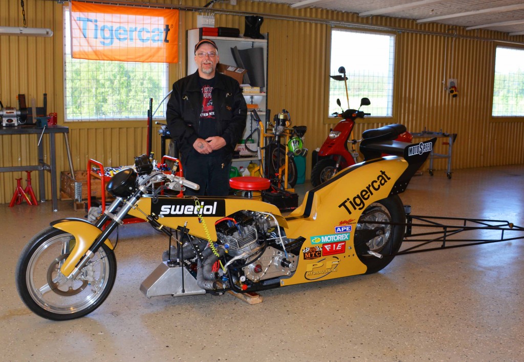 Mats debout à côté de son dragster dans un atelier, avec un drapeau de Tigercat flottant à la fenêtre derrière lui.