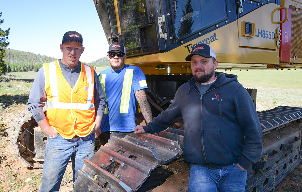 Three middle aged males stand in front of a Tigercat LH855D