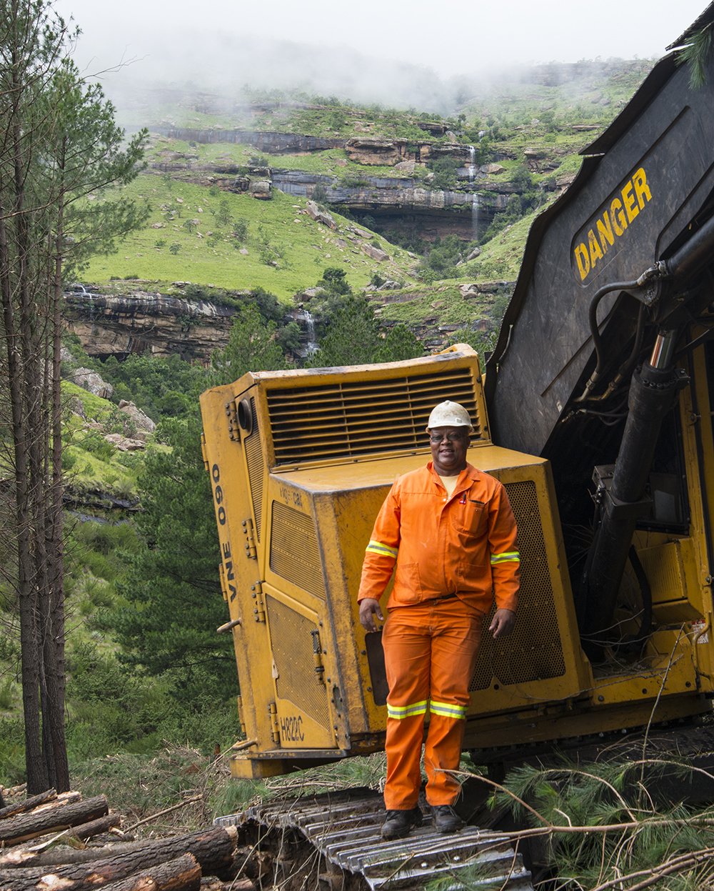 A Tigercat operator stands proud and smiling on the tracks of his machine in front beautiful hilly green landscape.