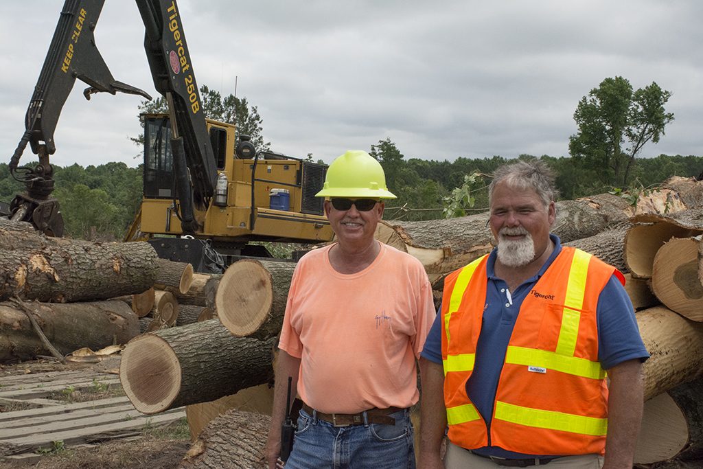 Two men stand smiling at a log site, a pile of logs behind them with a Tigercat 250B Loader in the background. 