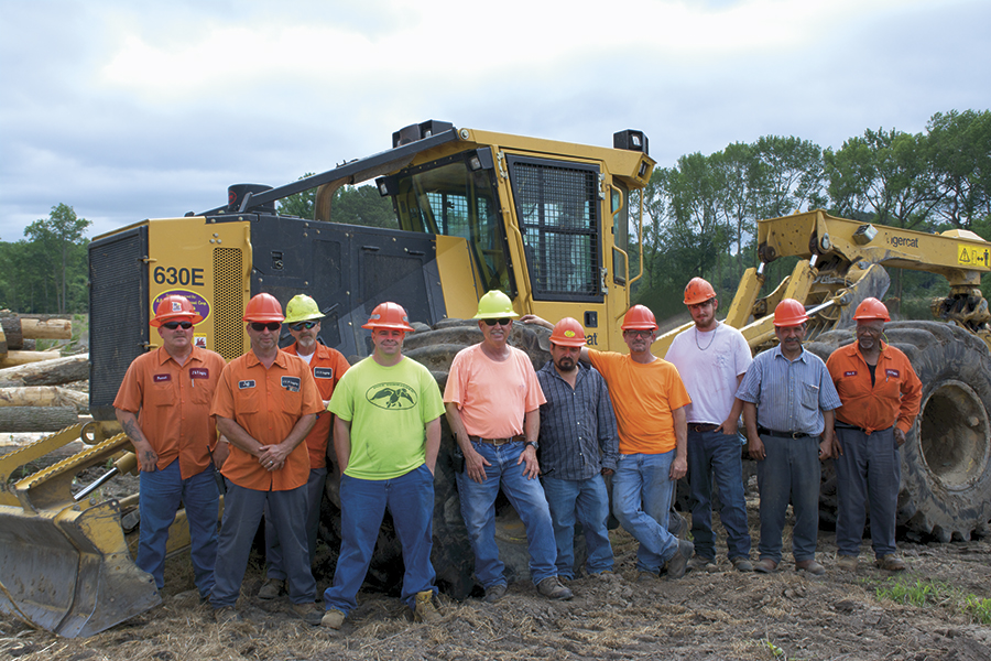The men of J & R Logging stand as a group smiling in front of a Tigercat 630E skidder. 