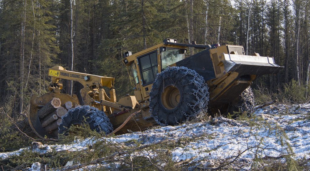The 4-wheel 630E pulling out of a steep gully with a full grapple of wood. Chains line the tires for extra traction on the snowy ground.