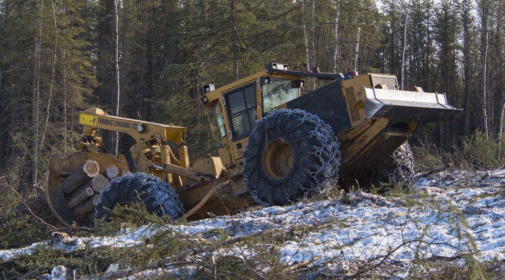 The 4-wheel 630E pulling out of a steep gully with a full grapple of wood. Chains line the tires for extra traction on the snowy ground. 