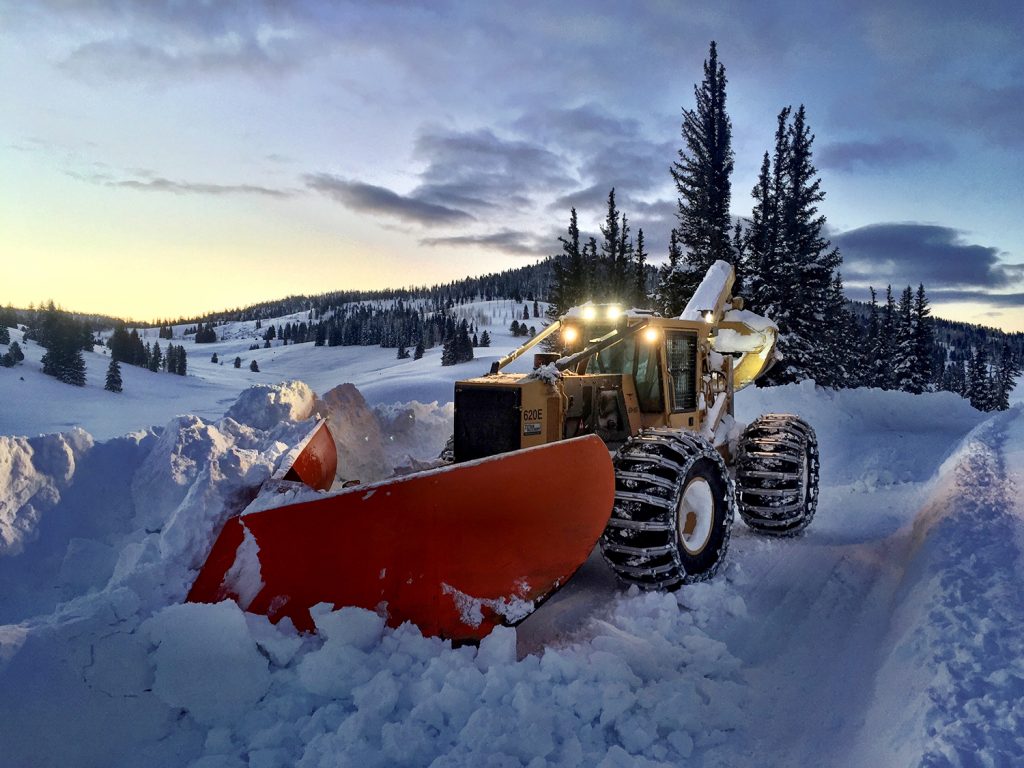 Трелевочный трактор Tigercat 620E, преобразованный в плужный снегоочиститель, очищает снег во время заката солнца в живописном окружении.