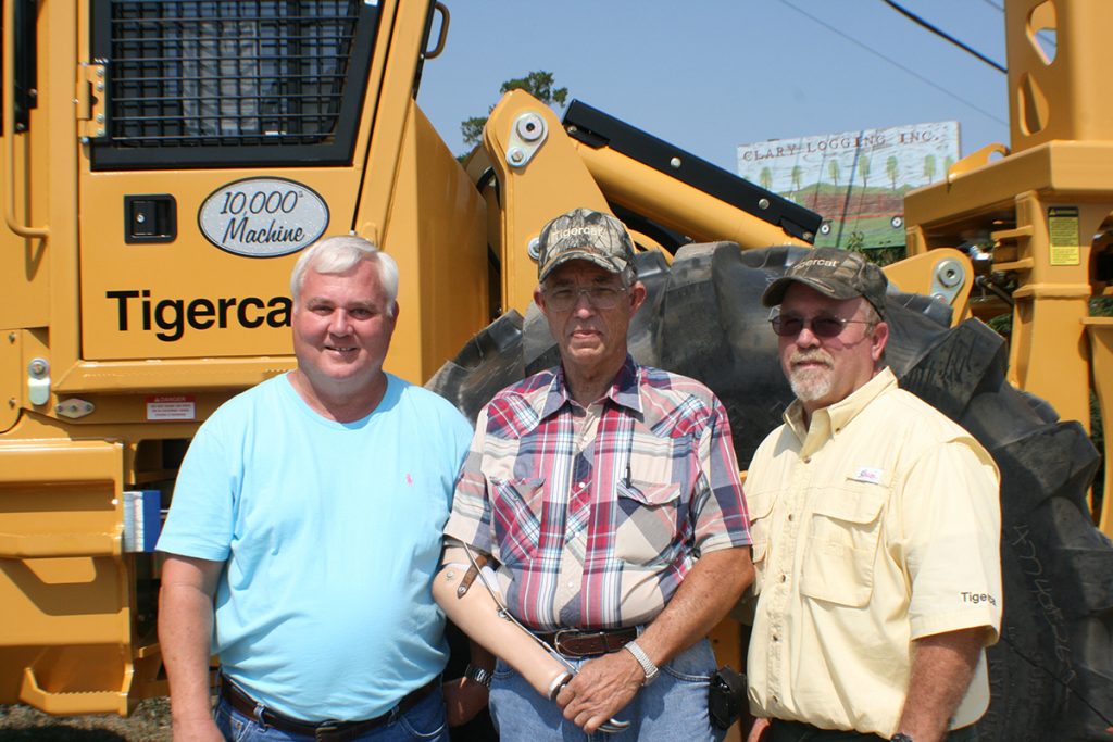 3 män står stolta och leende framför Tigercats 10 000:e tillverkade maskin. På en skylt i bakgrunden står det "Clary Logging Inc."