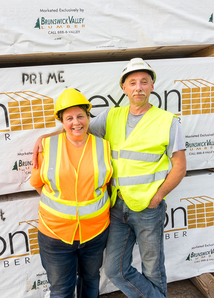 Kevin Sexton debout et passant son bras autour de sa femme, Susan Sexton, devant le bois de marque Sexton Lumber. Tout deux portent des gilets et des casques de sécurité.