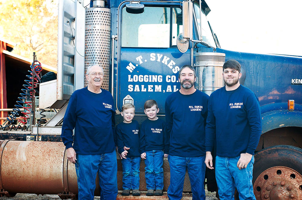 Four generations of Sykes loggers. (L-R) Bill, Cooper, Cam, Marvin and Colton stand in matching longsleeve shirts and jeans in front of a M.T. Sykes Logging transport truck.