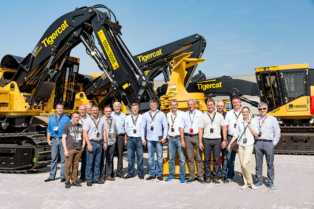 Визит группы «Илим» на завод Tigercat в Канаде; группа из 16 человек стоят вместе в окружении машин Tigercat на заводе Tigercat в Париже.