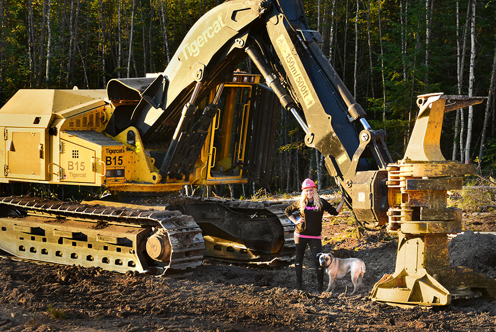 Loggergirl Hannah Dehoog with her pit bull Phoenix standing under the boom of her track feller buncher.