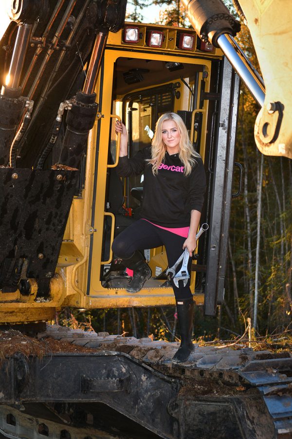 Passionate forestry equipment female operator, Hannah Dehoog of Smithers, British Columbia standing in the front door of her cab with wrenches in her hand wearing a Tigercat sweater.