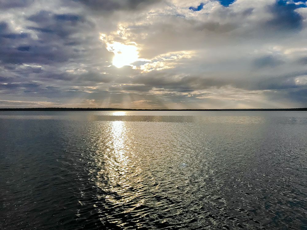 A lakescape, sun rays pierce from behind clouds, extending from the sky to the water. 