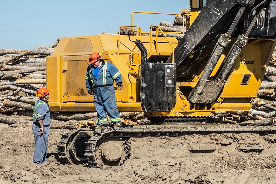 A man stands on the tracks of a Tigercat 880D logger looking down at another man on a job site. 