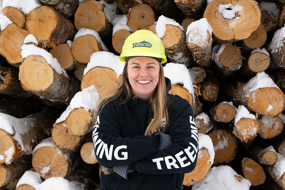 Whitney Souers stands with her arms crossed in front of a pile of logs. 